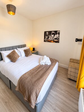 A neatly made bed with white linens and a brown throw blanket, a bedside table with a lamp, a wall-mounted power outlet, and a framed picture of a cityscape. - Newly Redecorated 1 bed flat ! (Weston-Super-Mare)