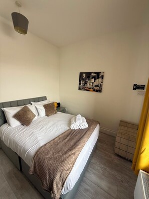 A hotel room with a bed, a nightstand, a lamp, a painting, and a window with curtains. - Newly Redecorated 1 bed flat ! (Weston-Super-Mare)