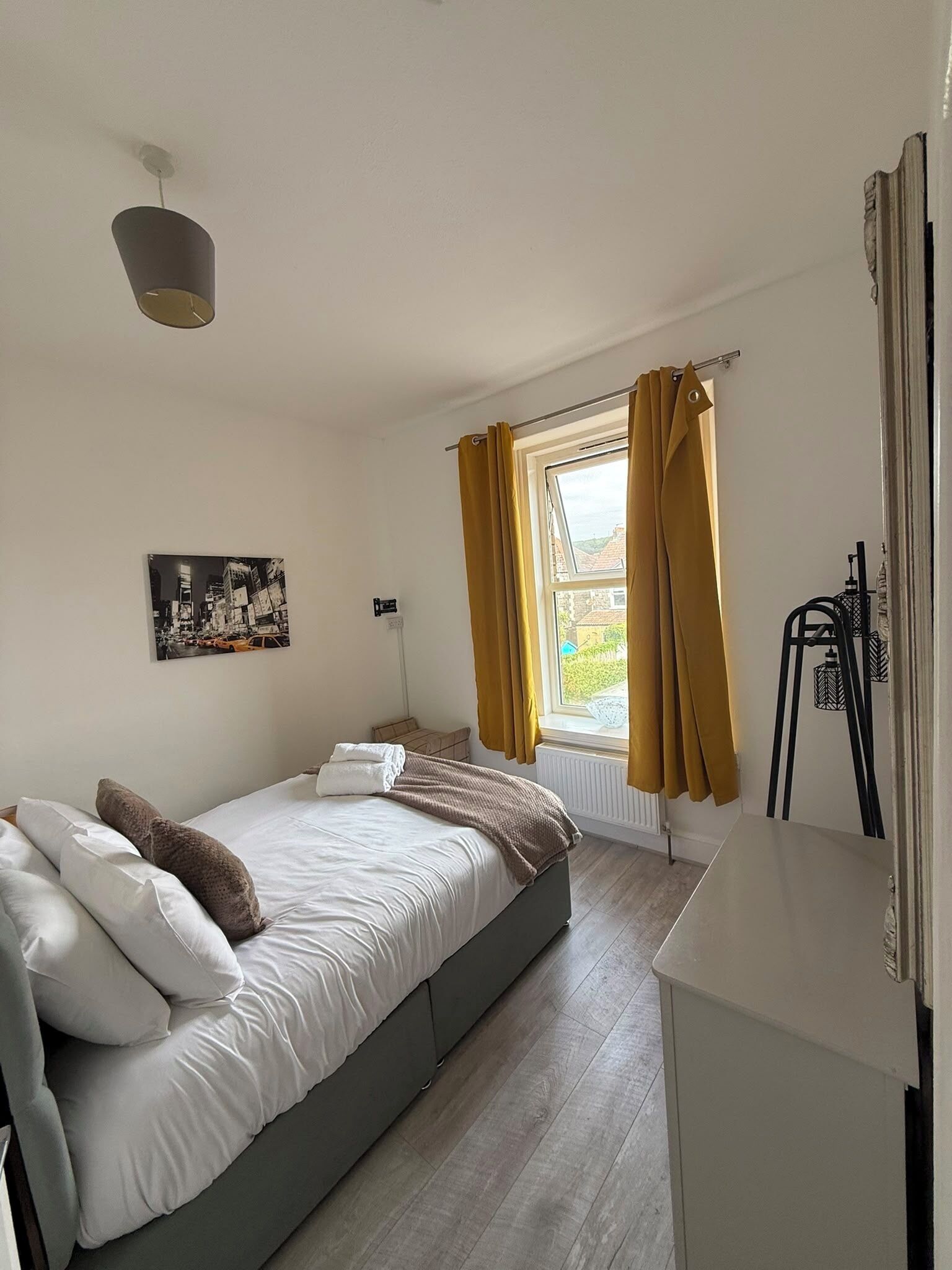 Quarto com uma cama de solteiro, janela com cortinas, radiador e um quadro na parede.