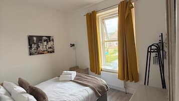 Quarto com uma cama de solteiro, janela com cortinas, radiador e um quadro na parede.
