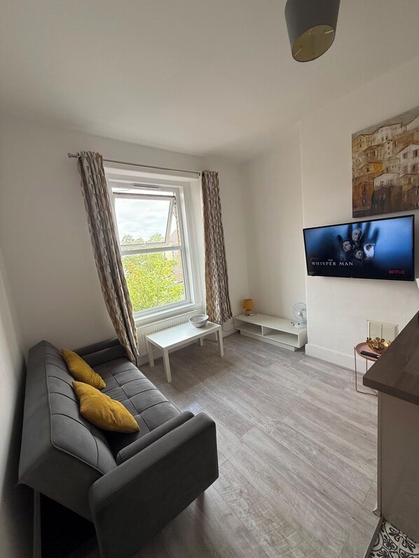 A modern living room with a grey sofa, a small white table, a TV mounted on the wall, and a window with curtains. - Newly Redecorated 1 bed flat ! (Weston-Super-Mare)