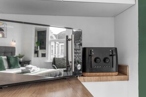 Una habitación con un espejo grande que refleja una cama y una ventana, una caja fuerte negra sobre un estante de madera, y una pared blanca.