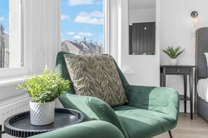 Una habitación con un sillón verde, una planta en maceta y vistas a edificios a través de una ventana.