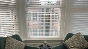 Een kamer met twee groene fauteuils, een kleine ronde tafel met een plant, en uitzicht op gebouwen door grote ramen met zonwering.