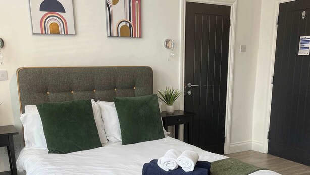 Een netjes opgemaakt bed met een grijs gestoffeerd hoofdbord, een groene plaid en opgerolde handdoeken. Twee aan de muur bevestigde kunstwerken, een donkere deur met de tekst 'THE LOO', en een kleine potplant op een nachtkastje.