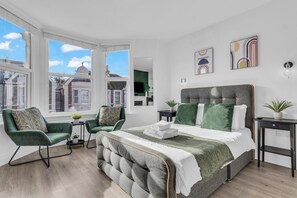 A modern bedroom with a large bed, two green armchairs, a small side table, and a view of the city through large windows. - 1st Floor 1 Bed Studio (Weston-Super-Mare)