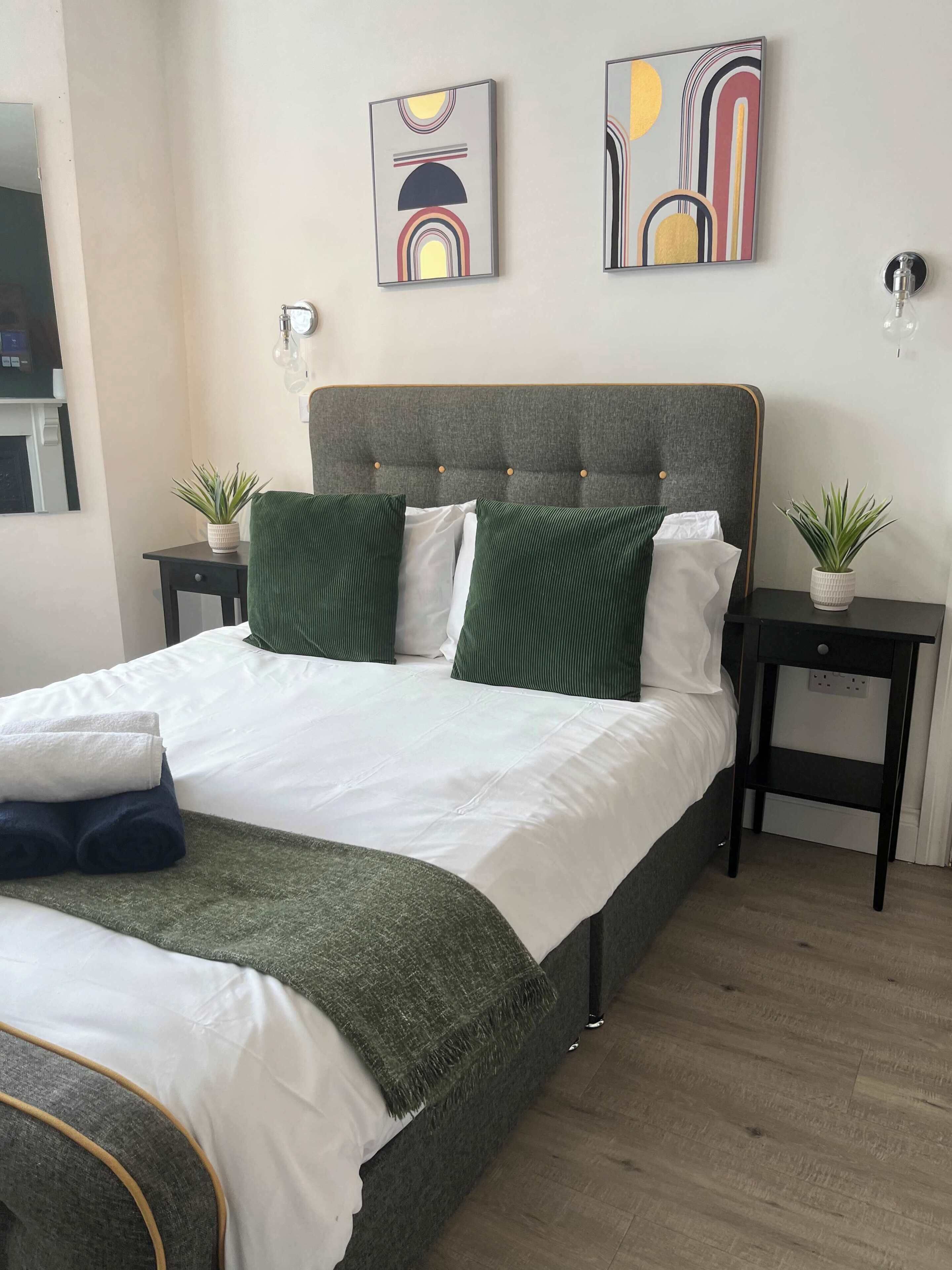 A modern bedroom with a grey upholstered headboard, a large bed with green and white bedding, two bedside tables with potted plants, and abstract wall art.