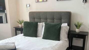 A modern bedroom with a grey upholstered headboard, a large bed with green and white bedding, two bedside tables with potted plants, and abstract wall art.