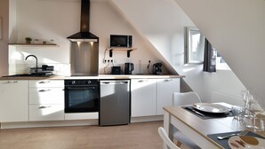 A modern kitchen with white cabinets, a dishwasher, a stove, and a microwave. There is a dining table with chairs and a window providing natural light. - Les Trois Rois 5 - Centre Avranches (Avranches)