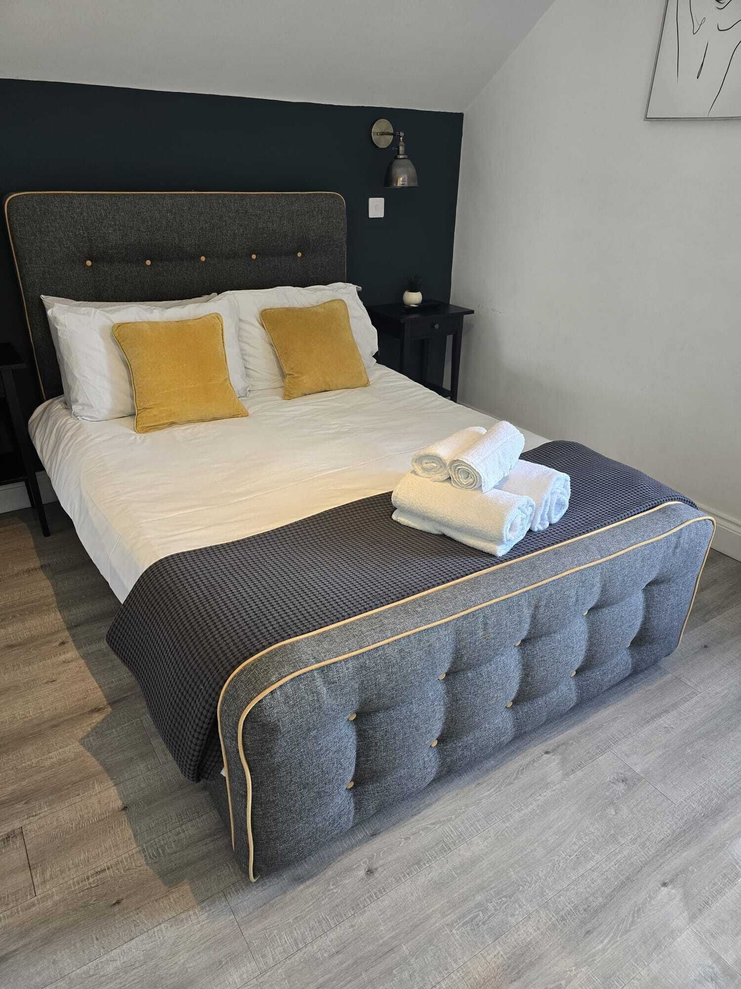 A neatly made bed with white and mustard pillows, a grey upholstered bench, and a small bedside table with a lamp.
