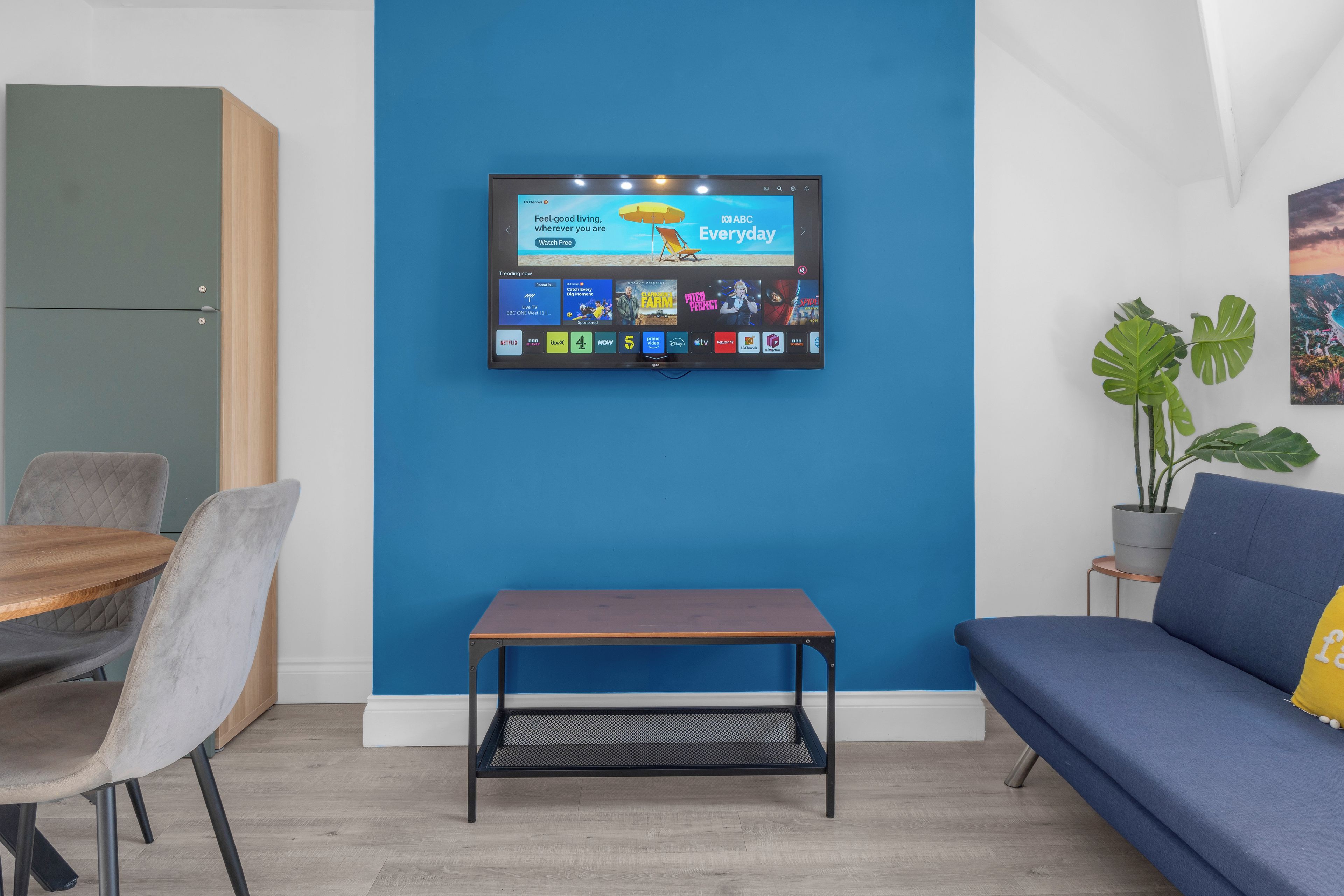 Un salon moderne avec un téléviseur à écran plat fixé sur un mur bleu, une table basse en bois, un canapé bleu et un coin repas avec des chaises.