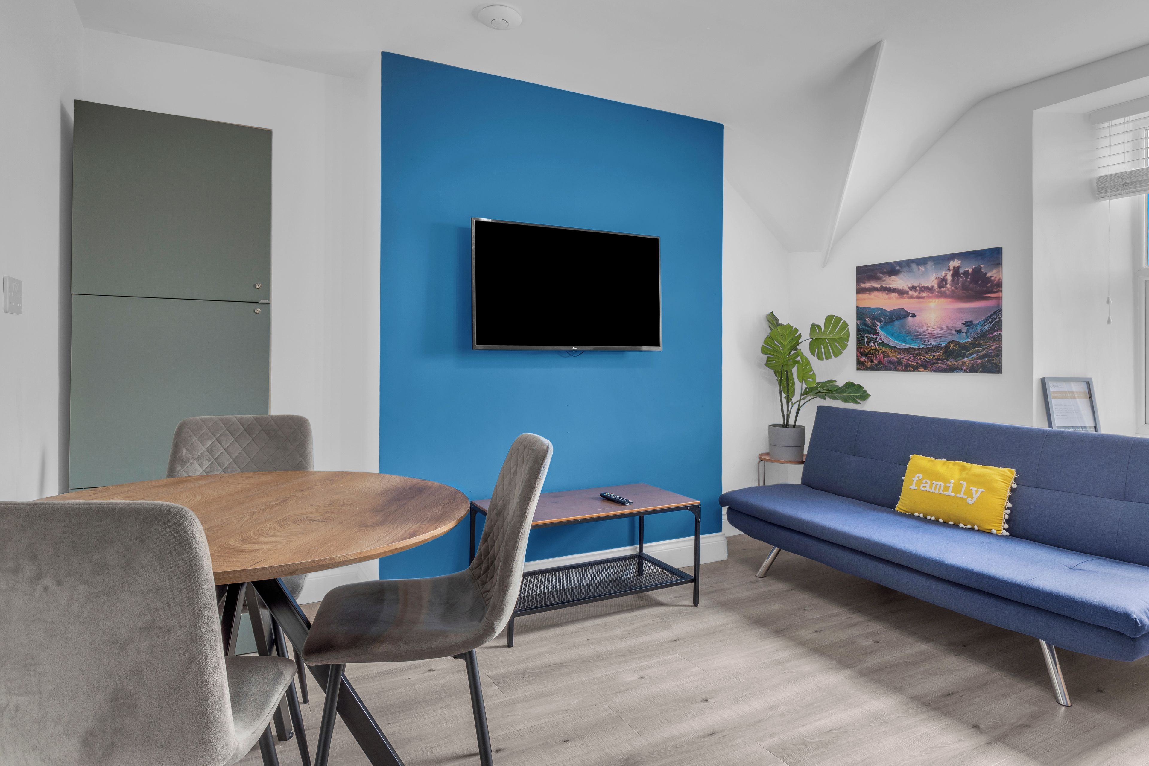 A modern living room with a blue accent wall, a flat-screen TV, a sofa with a 'family' pillow, a dining table with chairs, and a potted plant.