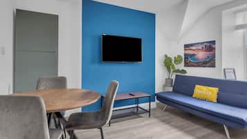 A modern living room with a blue accent wall, a flat-screen TV, a sofa with a 'family' pillow, a dining table with chairs, and a potted plant.