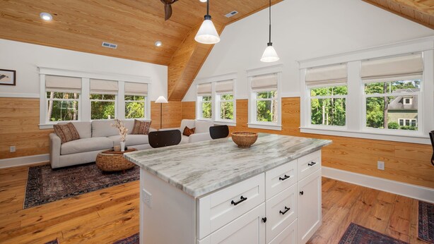 A spacious living room with a high ceiling, wooden beams, and large windows offering a view of greenery. - Award-Winner “The Carriage House”Gym+Pool Access (Powells Point)