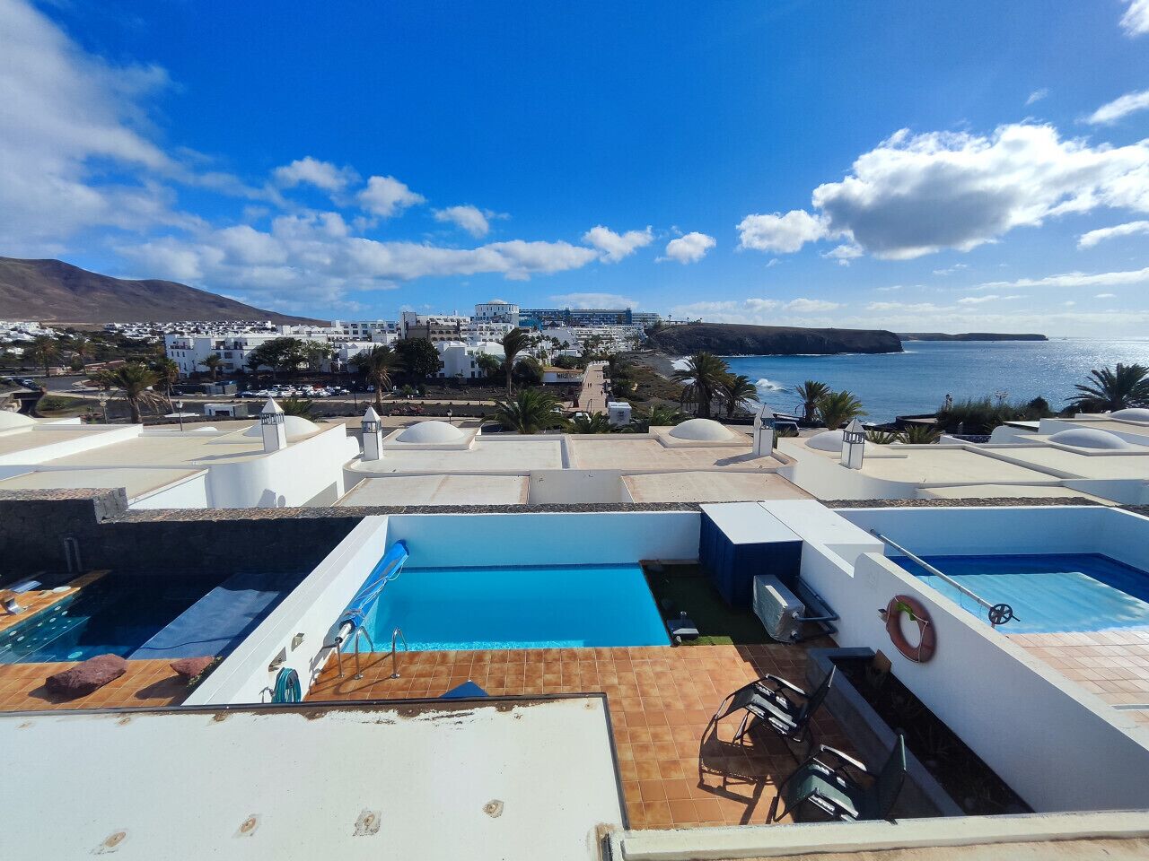 Un área de piscina en la azotea con varias piscinas, un salvavidas y vista al mar y edificios distantes.