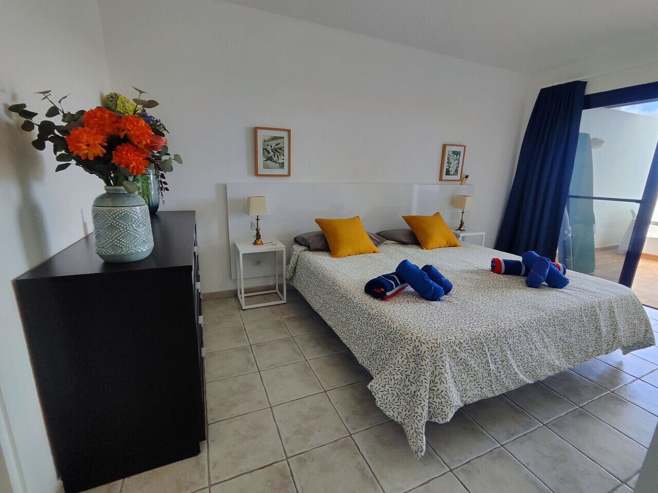 A bedroom with a bed, two bedside tables, a nightstand, a vase with flowers, and a window with curtains.