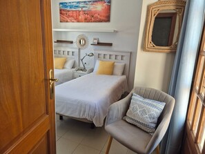 A hotel room with two beds, a chair, a nightstand, a lamp, and a painting on the wall. - Villa Adel: Oasis de paz con piscina climatizada (Playa Blanca)