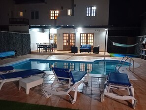 Una piscina con sillones de descanso, un muro de piedra y un edificio con ventanas iluminadas al fondo.