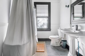 A bathroom with a shower curtain, a window with blinds, a toilet, a sink, and a trash can.