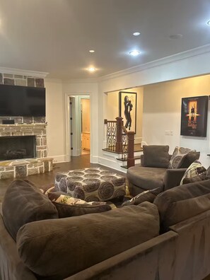 A living room with a stone fireplace, a large flat-screen TV, a staircase with a railing, and two posters on the wall. - Private Buckhead Estate Pool, Game Room, Custom Kitchen (Atlanta)