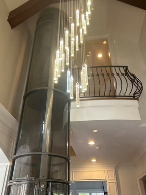 A modern interior with a glass elevator shaft, a decorative light fixture, and a balcony railing. - Private Buckhead Estate Pool, Game Room, Custom Kitchen (Atlanta)