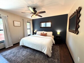 Una camera da letto con un letto, una ventola a soffitto, due lampade sui comodini, una finestra con tende e un quadro appeso al muro.
