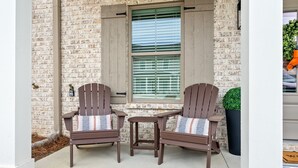 Eine Ziegelaußenwand mit zwei Adirondack-Stühlen und einem kleinen Tisch, ein Fenster mit Jalousien und einem Blumentopf.