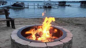 En rund stenanordning för en öppen eld, med flammor, omgiven av sand, nära ett vattendrag med båtar och en träbänk.