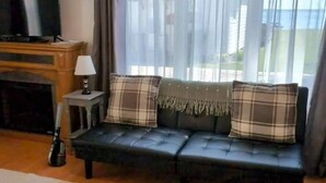Ett vardagsrum med en svart lädersoffa, en tv, en öppen spis och utsikt över naturen genom tunna, genomskinliga gardiner.