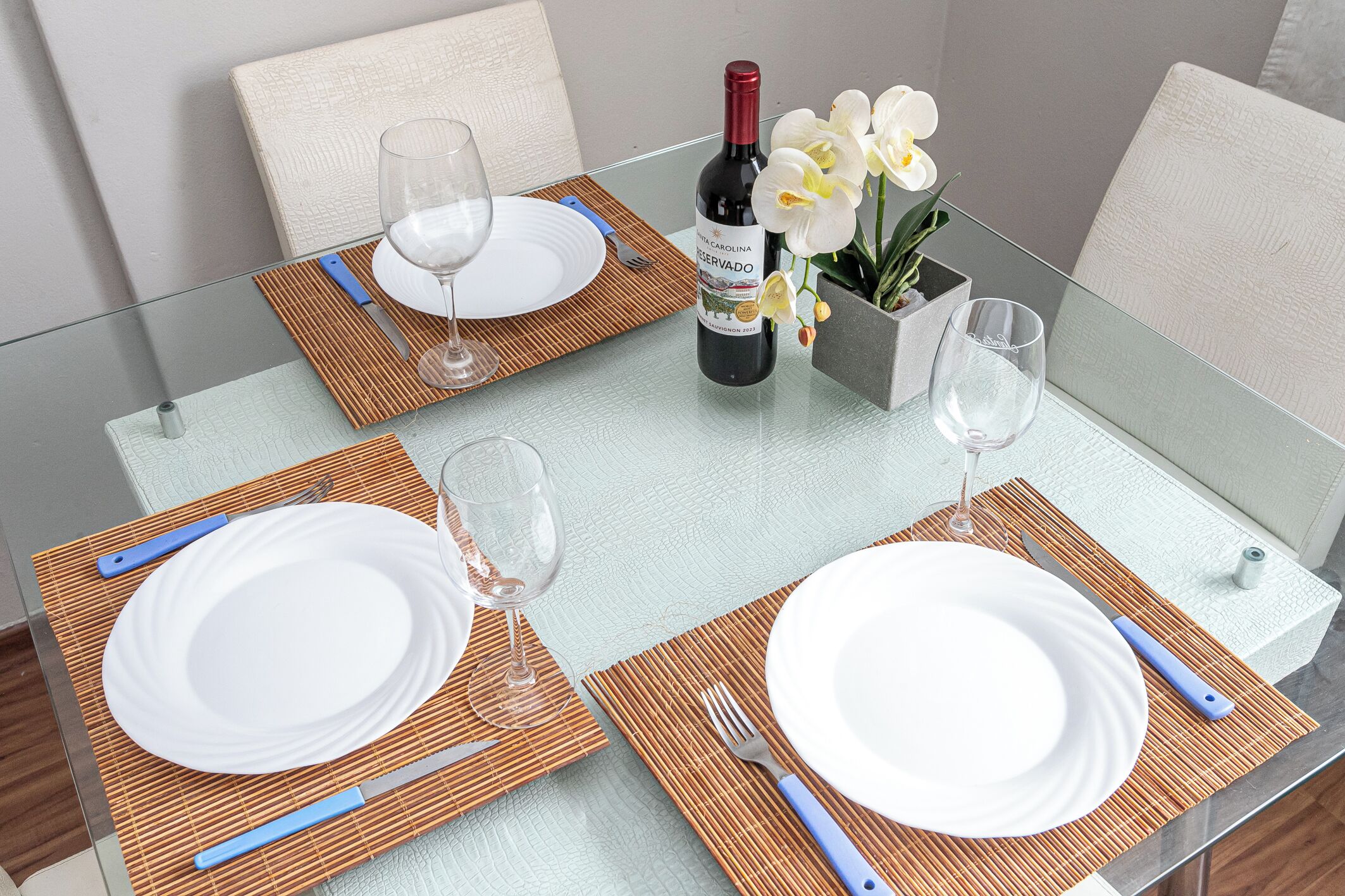 A dining table set with white plates, blue cutlery, and a bottle of wine.