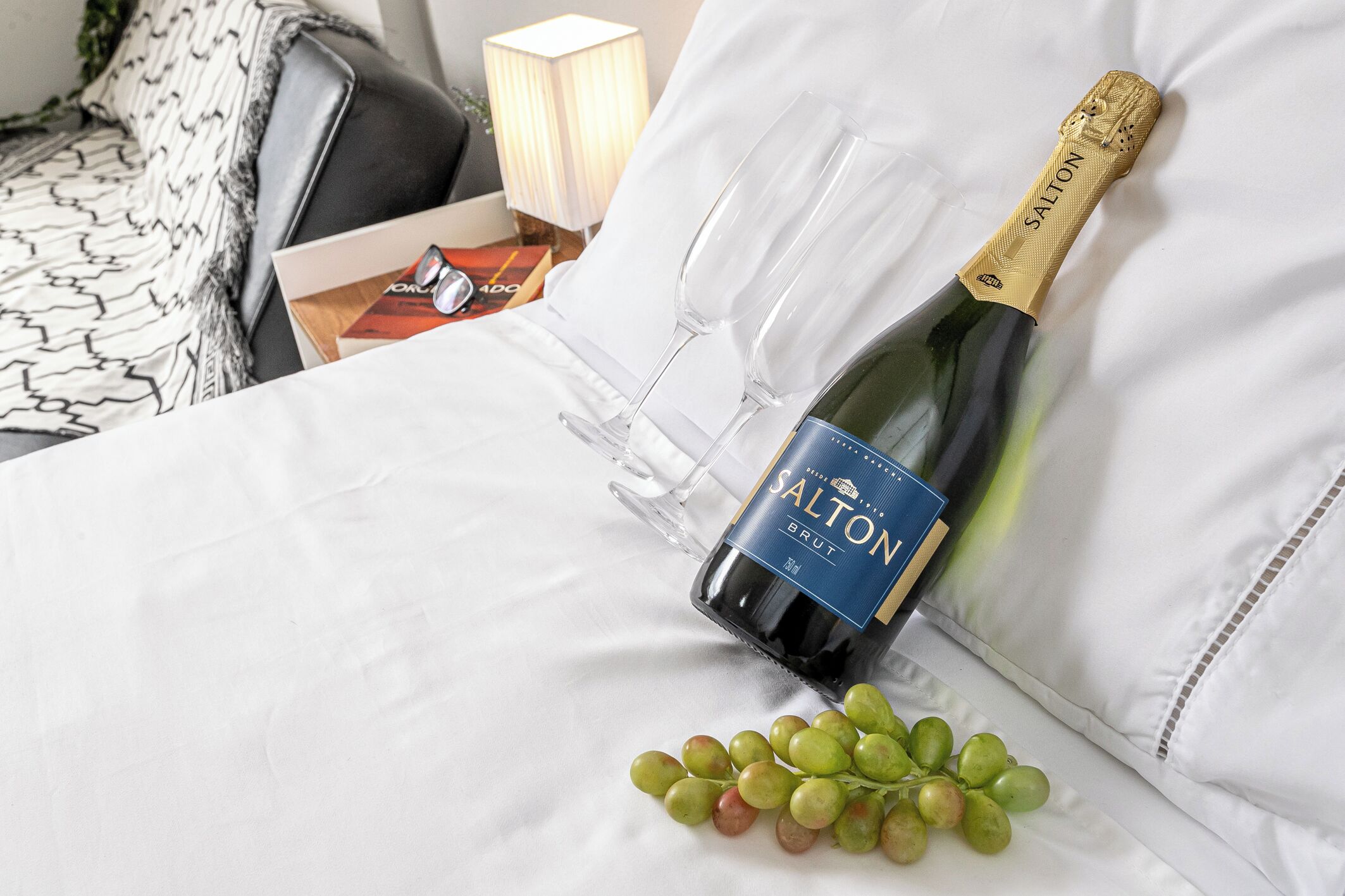 A bottle of Salton Brut champagne, a glass, and a bunch of grapes on a bed.