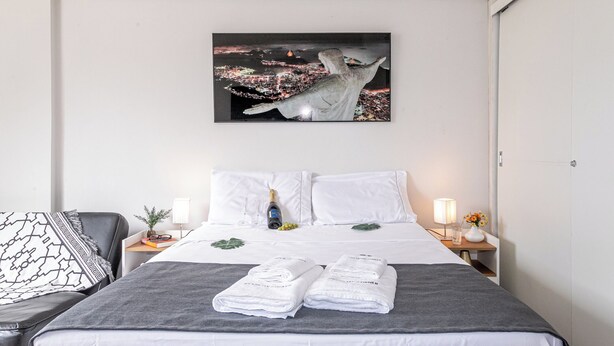 A hotel room with a bed, a nightstand with a lamp, a framed picture of a cityscape, and a window with a view of the city. - Incredible view of Cristo Redentor (Rio de Janeiro)