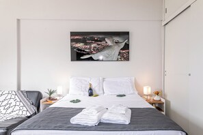 A hotel room with a bed, a nightstand with a lamp, a framed picture of a cityscape, and a window with a view of the city. - Incredible view of Cristo Redentor (Rio de Janeiro)