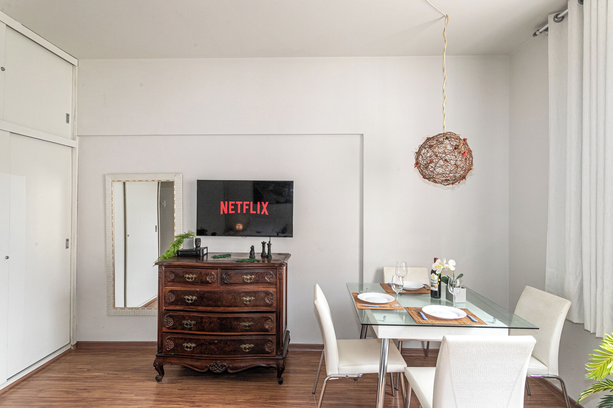 A modern dining area with a glass table, white chairs, a wooden dresser, and a television displaying 'NETFLIX'.