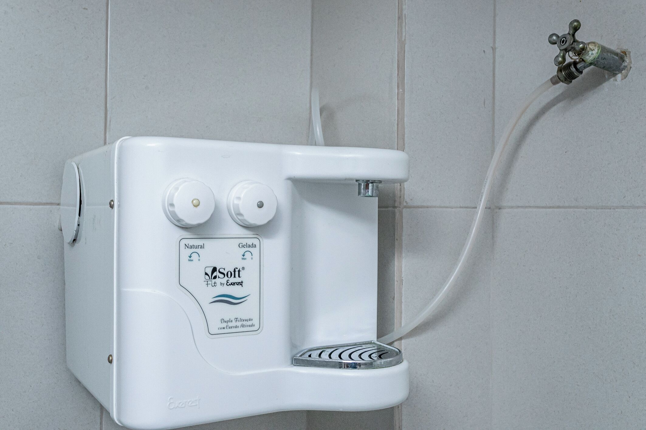 A white water softener unit with two control knobs and a drain pump, mounted on a tiled wall.