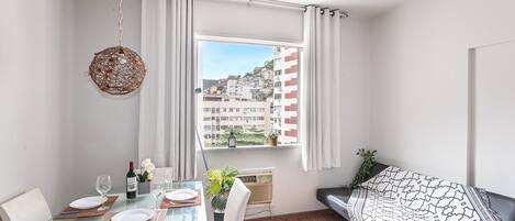 A modern living room with a glass dining table, white chairs, a black sofa, and a view of a cityscape through a large window.