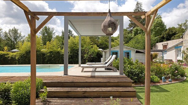 Una pérgola de madera con una lámpara colgante, una piscina y una casa con puerta azul al fondo.