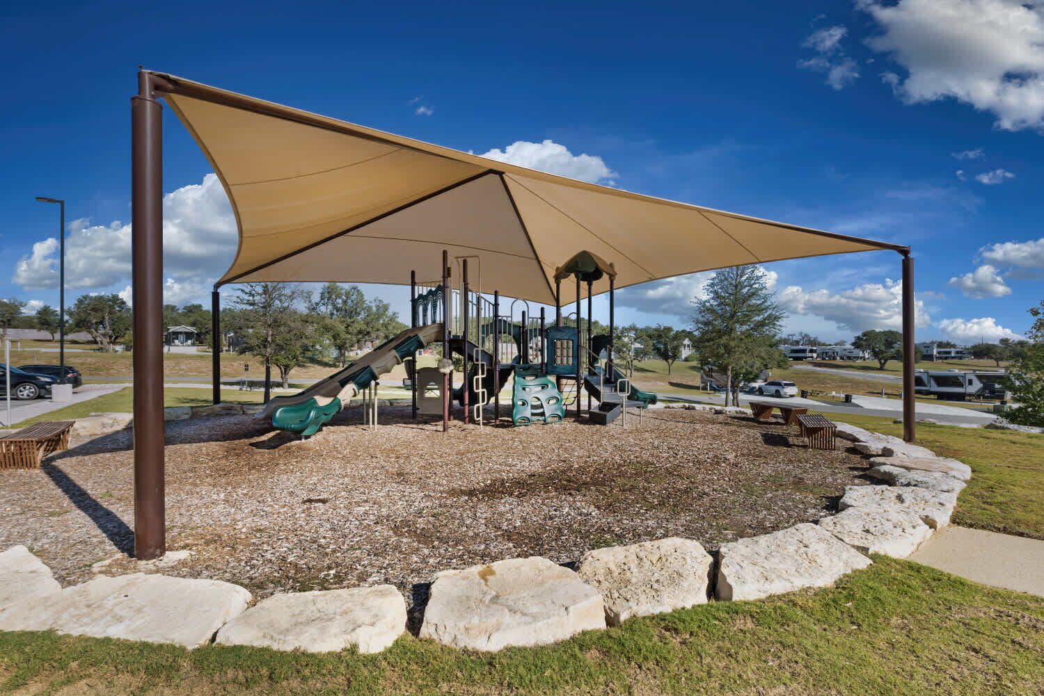 A playground with a slide and climbing structures, surrounded by a shaded area and a gravel path.