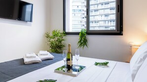 A hotel room with a bed, a bedside table with a bottle of champagne and two glasses, a TV mounted on the wall, and a window with a view of a building.