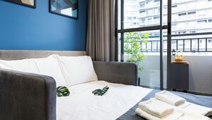 A hotel room with a bed, a grey sofa, a window with a city view, and a small table with a plant and a bottle.