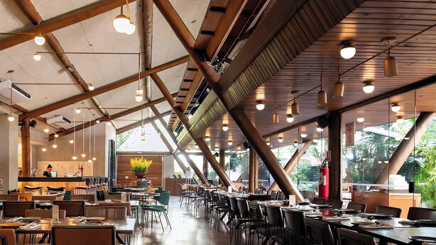A spacious restaurant with high ceilings, exposed beams, and large windows offering views of greenery. Tables and chairs are neatly arranged, and a bar area is visible in the background.