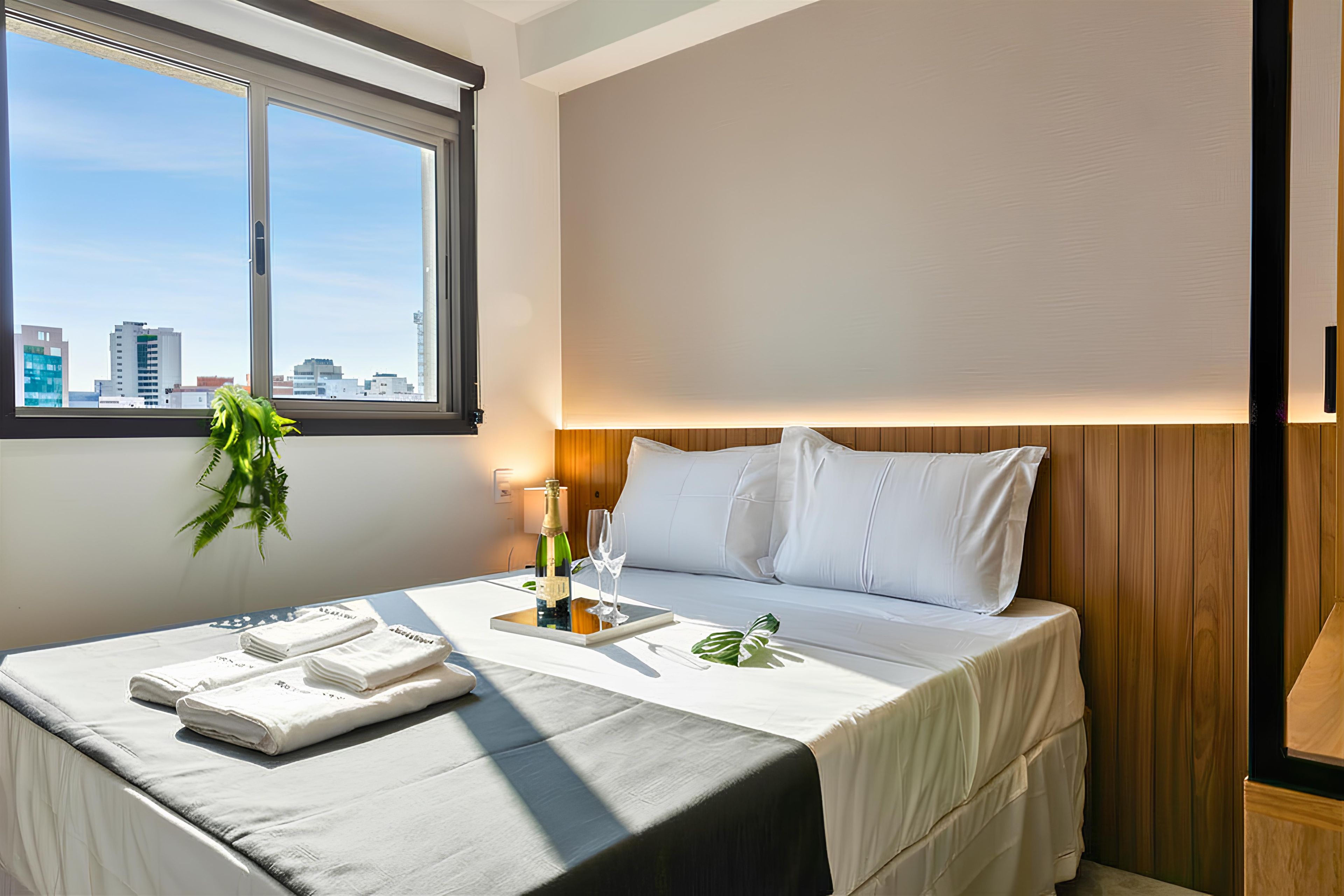 Habitación de hotel con cama, mesita de noche con botella y vasos, ventana con vista a la ciudad y una planta.