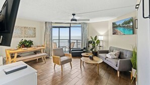 Ein modernes Wohnzimmer mit einer Couch, Sesseln, einem Couchtisch und einem Essbereich. Ein Balkon bietet Blick aufs Meer.