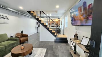 A modern living room with a green sofa, a brown armchair, and a wooden coffee table. A staircase leads to a kitchen area with a dining table and chairs.