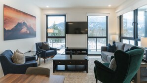 A modern living room with a large TV, comfortable seating, and large windows offering a view of the outdoors. - The Luxury Basecamp in Mackenzie Village (Revelstoke)