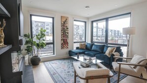 A modern living room with a blue sofa, a coffee table, and a view of the city through large windows. - The Big Lohbow - Ski Haus in Mackenzie Village (Revelstoke)