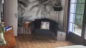 Et soveværelse med en mørk sofa, en hvid, blød måtte, en seng med lyseblåt sengetæppe, et træsidebord, et lille sidebord, en lampe og en væg med et palmearbejde.