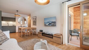 A modern living room with a sofa, coffee table, and dining area. There is a fireplace, a large window, and a sliding glass door leading to an outdoor deck. - Kandahar – Warm Alpine Retreat in Les Gets (Les Gets)