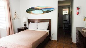 A bedroom with a bed, a nightstand, a lamp, a painting of a beach scene, and a fire extinguisher. - Feel safe and rest in our cozy 1-bedroom studio with AC (Manatí)