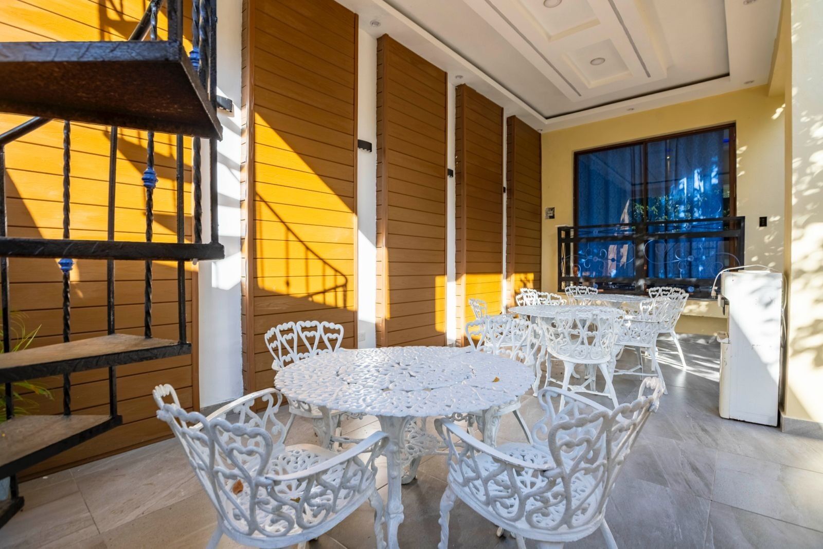 A sunlit outdoor patio with white wrought iron tables and chairs, wooden panel walls, and a view of the city through large windows.